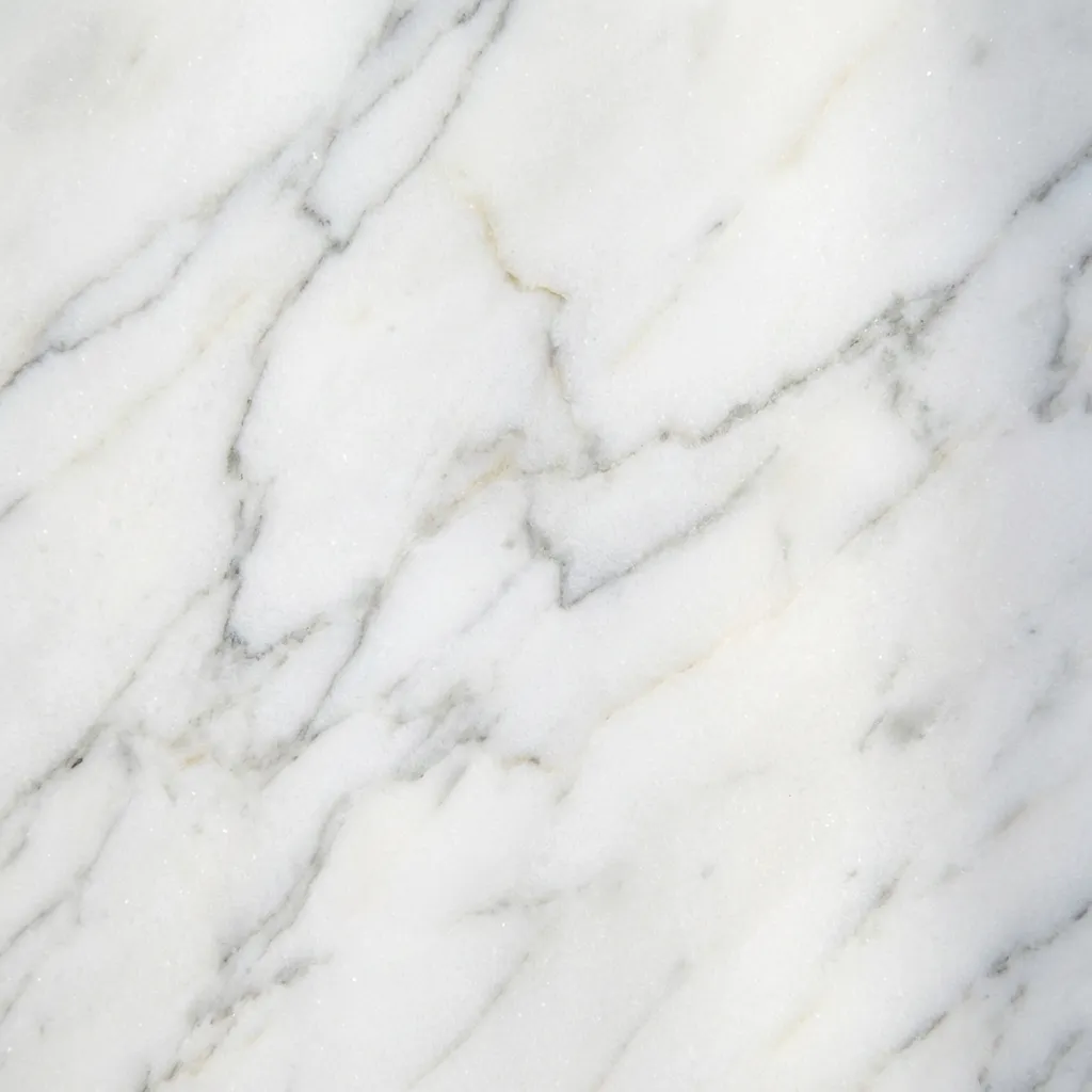 磨き上げられた白い大理石の、その見事な自然な質感をクローズアップで捉えた画像です。滑らかで清らかな表面には、濃淡のあるエレガントなグレーの脈理が斜めに交差し、温かみのあるベージュやゴールドの繊細な色合いが控えめに添えられています。これにより、豪華なインテリアやミニマリストデザインに最適な、洗練された時代を超越した美学が生まれています。