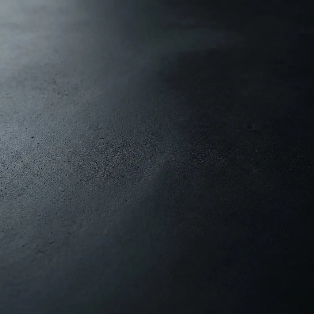 ローアングルで捉えられた、コンクリートかアスファルトのような粗い質感の暗いグレーの表面のクローズアップ写真です。柔らかい光が手前を穏やかに照らし、そのざらついたディテールを際立たせ、奥に行くほど深い影へと溶け込み、ミニマルで静謐な雰囲気を作り出しています。