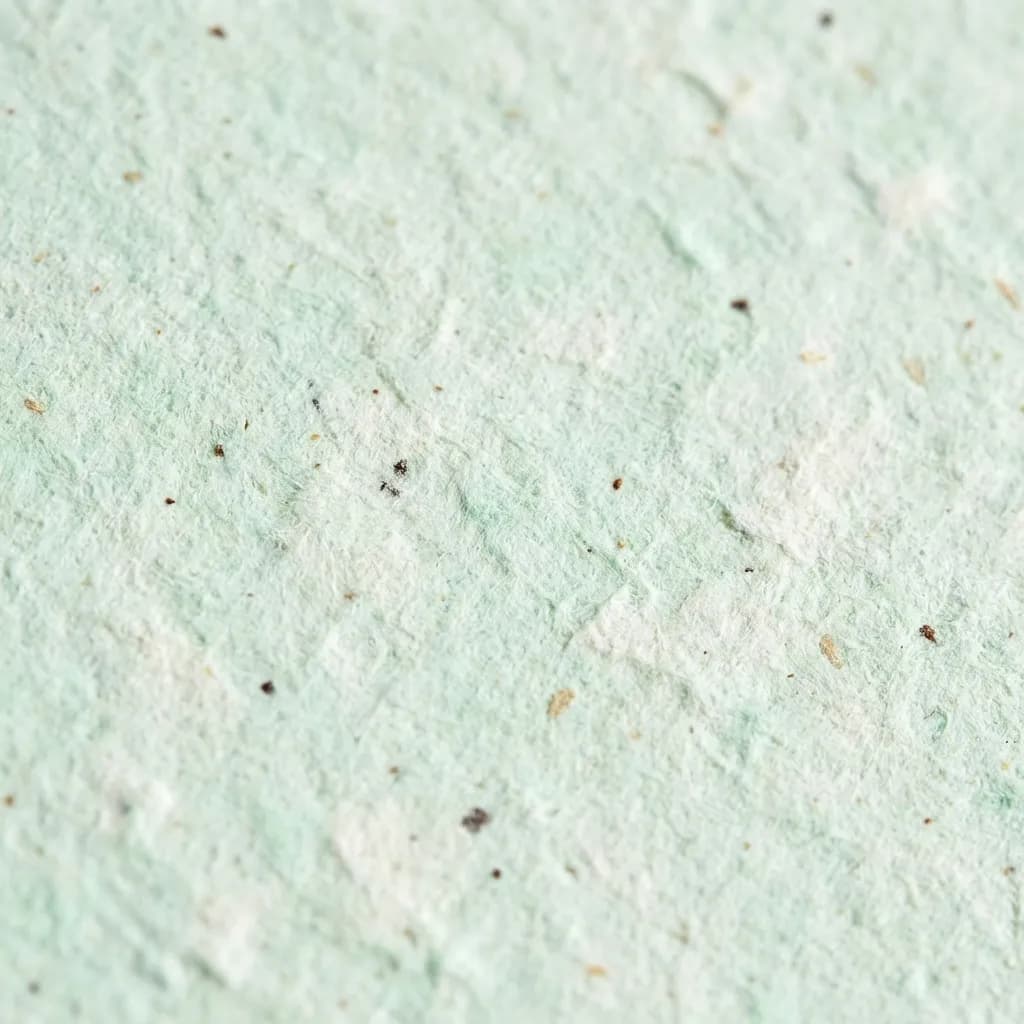 柔らかく落ち着いたミントグリーンを基調とした、手漉き紙のテクスチャをクローズアップで捉えた画像です。紙の表面は不均一で繊維質がはっきりと見え、小さな濃い茶色の斑点や薄いベージュの破片が埋め込まれており、オーガニックで素朴な雰囲気を醸し出しています。この魅力的な質感と自然な色合いの紙は、環境に優しく、職人の技を感じさせる美しさを持っています。